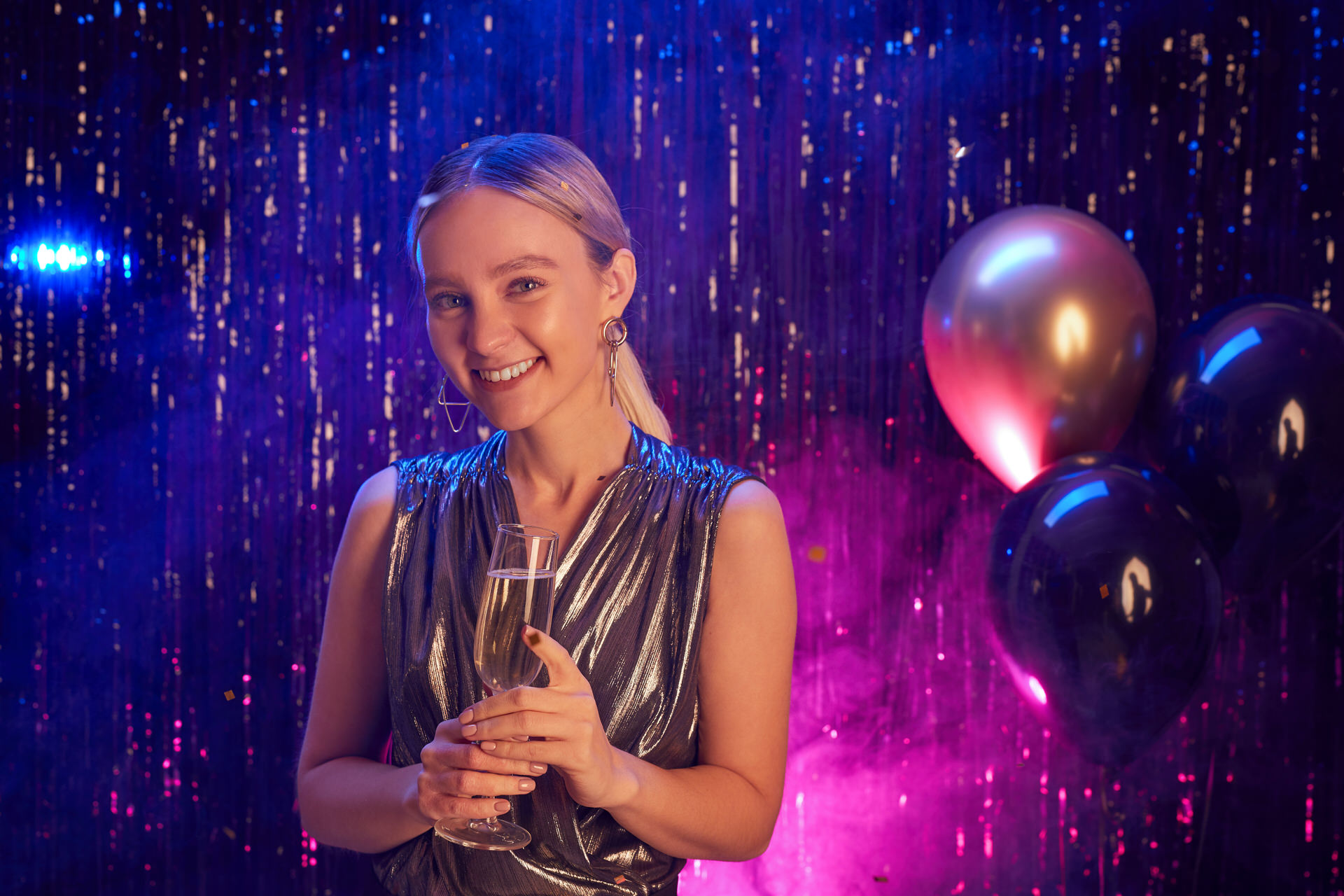 Portrait of blonde young woman holding champagne glass and smiling at camera while enjoying party in nightclub, copy space